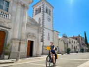 Radfahrerin mit gelben Taschen fährt über sonnigen Platz in Gradisca d'Isonzo vorbei an weißer Kirche mit Glockenturm unter blauem Himmel.