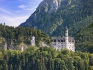 Neuschwanstein Castle with white towers nestled in dense forest below a rocky mountain peak in Bavaria, Germany, under blue sky.