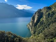Panoramic view from Strada del Ponale showing steep cliffs covered with vegetation overlooking the blue waters of Lake Garda with mountains.