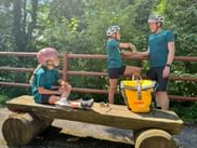 Family of three in matching teal cycling gear resting on wooden bench. Child in pink helmet sits on bench, parents stand behind with yellow Eurobike bag.
