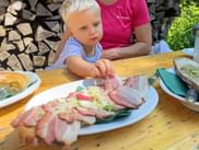 Blonde child reaching for sliced bacon bread on a plate at an outdoor wooden table, with an adult in pink shirt and stacked firewood in background.
