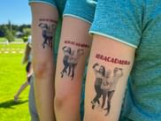 Three people wearing turquoise shirts display matching temporary tattoos reading 'ABRACADABRA' on their upper arms at Grabenseelauf 2026.