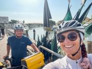Zwei Radfahrer mit Helmen machen ein Selfie am Hafen von Caorle. Traditionelle Fischerboote mit grünen Netzen dahinter, Kanal und Gebäude im Hintergrund.