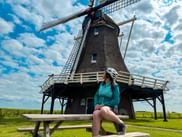Radfahrerin in türkisfarbener Jacke und Helm sitzt auf Picknicktischbank vor traditioneller holländischer Windmühle in Harderwijk unter bewölktem Himmel.