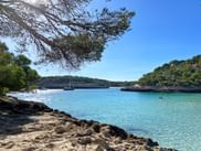 Malerische Bucht in Porto Colom, Mallorca mit türkisem Wasser, felsiger Küste, Pinien, Segelboot und grünen Hügeln unter blauem Himmel.