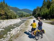 Two cyclists with touring bikes and panniers on a path beside Krimmler Ache river in Krimml, with mountains and forests in the background.