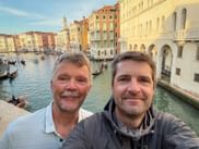 Two men taking a selfie on a bridge over Venice's Canal Grande with colorful historic buildings, gondolas, and a church tower in the background.