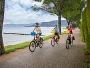 Drei Radfahrer mit Packtaschen fahren auf einem baumgesäumten Uferweg am Gardasee. Berge sind über dem ruhigen blauen Wasser sichtbar.