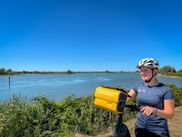 Lächelnde Radfahrerin mit weißem Helm hält gelbe wasserdichte Lenkertasche am Canale di Primero in Friaul, mit ruhigem Wasser und blauem Himmel.