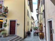 Schmale Kopfsteinpflastergasse in Udine mit gelben Gebäuden, Holztüren und Menschen an Tischen im Freien unter blauem Himmel.
