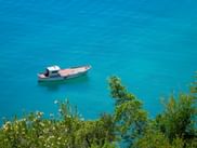 Small fishing boat floating on crystal-clear turquoise water near Rab island, Croatia. Green vegetation is visible in the foreground.