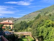 Stone arch bridge spanning a green valley in Tuscany with a house, hills covered in olive groves, and blue sky above.