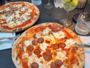 Zwei italienische Pizzen mit Salami und Mozzarella auf einem Restauranttisch in Caorle, begleitet von Weingläsern und Wasser.