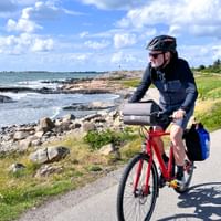 Radfahrer mit Helm auf rotem Fahrrad auf Küstenweg entlang der schwedischen Westküste bei Varberg, mit felsiger Küste und blauem Meer.
