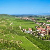 Luftaufnahme des Dorfes Ammerschwihr im Südelsass, umgeben von terrassierten Weinbergen unter blauem Himmel mit fernen Bergen.