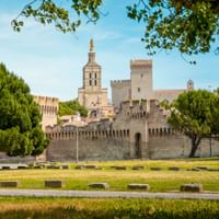 Blick auf Avignons befestigte Stadtmauern mit Türmen und dem Papstpalast dahinter, eingerahmt von grünen Bäumen und Rasenfläche in der Provence.