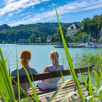 Two women sitting on a bench on a jetty in Weyer Bay with a view of Mattsee Castle