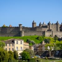 Mittelalterliche Festung Carcassonne mit Steinmauern und Türmen auf einem Hügel, umgeben von grünem Gras und Bäumen. Wohngebäude im Vordergrund.
