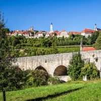 Historische Steinbrücke mit Bögen im Vordergrund, mittelalterliche Stadt Rothenburg ob der Tauber mit rotgedeckten Gebäuden und Kirchtürmen auf Hügel.