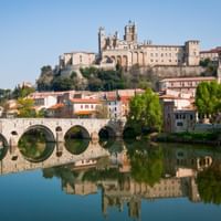 Stone bridge with multiple arches spanning a calm river in Béziers, with Cathedral Saint-Nazaire on the hilltop and reflections in the water.