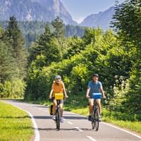 Zwei Radfahrer auf asphaltiertem Weg durch das Kanaltal bei Tarvis, umgeben von grünem Wald mit Berggipfeln im Hintergrund.