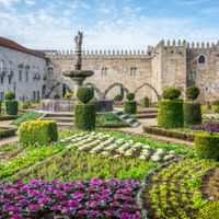 Jardim de Santa Barbara in Braga mit bunten Blumenbeeten, geschnittenen Hecken und einem Brunnen. Mittelalterliche Steingebäude im Hintergrund.