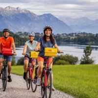 Drei Radfahrer auf einem Schotterweg am Riegsee in Bayern, mit grünen Wiesen und dramatischen Alpenbergen im Hintergrund.