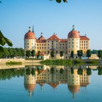 Schloss Moritzburg mit gelber Fassade und roten Kuppeln spiegelt sich perfekt im ruhigen Wasser, eingerahmt von grünem Laub unter blauem Himmel.