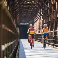 Zwei Radfahrer fahren durch eine historische Eisenbahnbrücke mit Holzfachwerk im Kanaltal auf dem Alpe-Adria-Radweg.