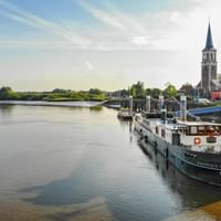 River cruise ship MS Magnifique 2 docked at a wooden pier in Sint-Amands. A church with tall spire is visible in the background under blue sky.