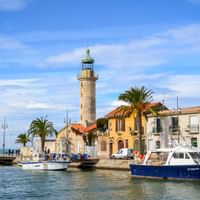 Steinerner Leuchtturm am Hafen von Le Grau-du-Roi mit Fischerbooten, mediterranen Häusern, Palmen und blauem Himmel in der Camargue, Provence.