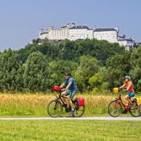 Drei Radfahrer mit gelben Taschen fahren auf einem Weg durch grüne Wiesen mit der Festung Hohensalzburg auf einem bewaldeten Hügel im Hintergrund.