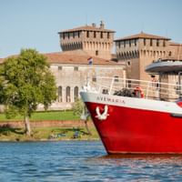 Rot-weißes Passagierschiff Ave Maria auf blauem Wasser vor historischen Festungsmauern und Türmen in Mantua mit grünen Bäumen am Ufer.
