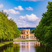 Yellow Orangerie palace in Kassel framed by symmetrical rows of green trees along a reflective canal under a blue sky with white clouds.