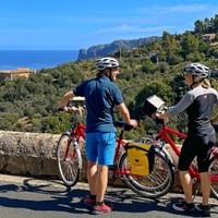 Two cyclists with red touring bikes and yellow panniers viewing the Mediterranean Sea from a coastal road in Mallorca.