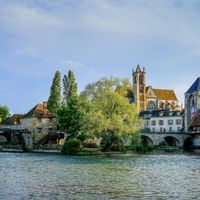 View of Moret-sur-Loing with stone bridge crossing the Loing river, historic church with tower, and traditional buildings under blue sky.