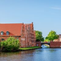 Rote Backstein-Salzspeicher am Kanal in Lübeck mit Kirchtürmen, Brücke und Ausflugsboot unter blauem Himmel.