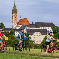 Three cyclists riding through a green meadow with Andechs Monastery visible on a hill in the background, featuring a baroque church tower.
