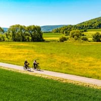 Two cyclists riding on a paved path through green and yellow meadows in the Weserbergland region with forested hills in the background.