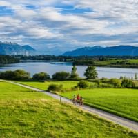 Two cyclists on a path through green meadows near Riegsee lake in Bavaria, with mountains in the background under a cloudy blue sky.