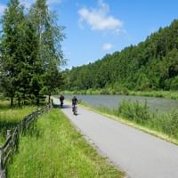 Cyclists on paved path along Dunajec River with wooden fence, green meadows, and forested hills under blue sky.
