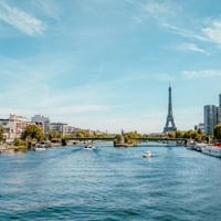 View along the Seine River in Paris with the Eiffel Tower in the distance, modern buildings on both banks, and boats on the water under a blue sky.