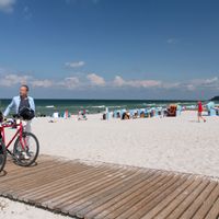 Two cyclists pushing their bikes on a wooden walkway on the beach, beach chairs in the background