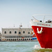 Rot-weißes Ave Maria Schiff in der Lagune von Venedig mit Markusturm und Dogenpalast im Hintergrund unter klarem blauen Himmel.
