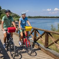Zwei Radfahrer mit Helmen und Packtaschen auf einer Holzstegbrücke in Grado, Friaul. Blaues Lagunenwasser und grüne Vegetation unter klarem Himmel.