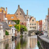 Scenic canal in Bruges lined with medieval brick houses with orange tiled roofs. A stone bridge crosses the calm water under a clear blue sky.