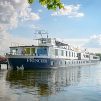 River cruise ship MS Princess moored at a wooden pier on calm water. The white vessel reflects in the water under a blue sky with clouds.