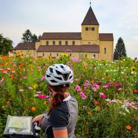 Eine Radfahrerin steht vor einer Blumenwiese und einer Kirche
