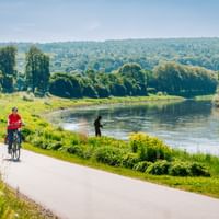 Zwei Radfahrer in roten Trikots auf asphaltiertem Weg entlang der Weser. Grüne Landschaft mit Bäumen und Hügeln im Hintergrund unter blauem Himmel.