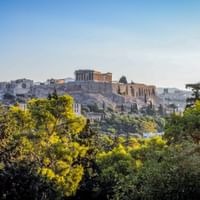 Blick auf die Akropolis mit dem Parthenon-Tempel in Athen, Griechenland. Antike Ruinen auf felsigem Hügel umgeben von grünen Bäumen unter klarem Himmel.